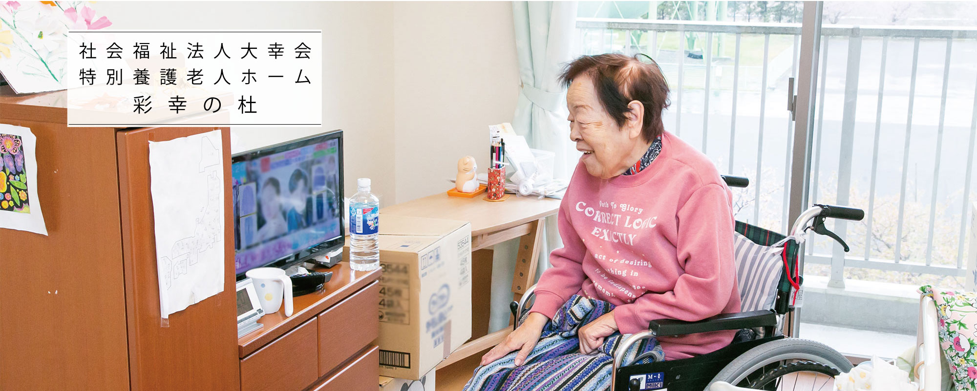 入居者がテレビを視聴している居室での日常風景。車椅子利用者が番組を楽しむ様子が映っている。